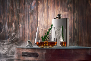 A crystal glass of whiskey with a sprig of rosemary and a metal flask, shrouded in clouds of smoke,...