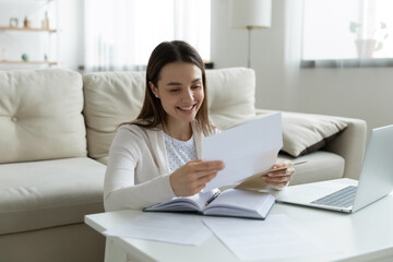 Smiling young woman reading unwrapped from paper envelope letter, excited by good news. Happy millennial girl checking post mail, received bank tax refund notification or loan approval at home.