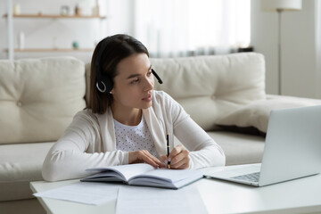 Pleasant young female PhD student in headphones sitting at coffee table in living room watching educational lecture, attending online courses webinar, consulting with supervisor about thesis writing.