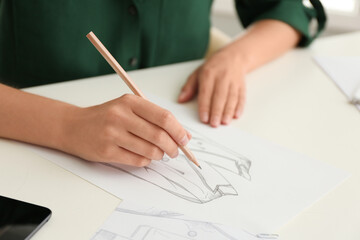 Woman drawing clothes with pencil on sheet of paper at white table, closeup