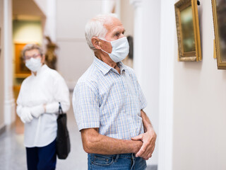 mature man in mask protecting against covid examines paintings on display in hall of art museum