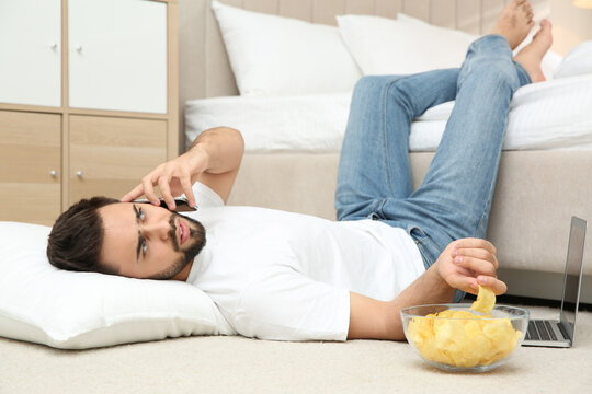 Lazy young man with laptop, smartphone and bowl of chips lying on floor at home