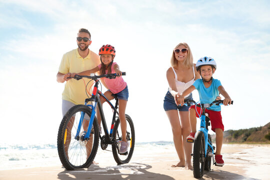 Happy parents teaching children to ride bicycles on sandy beach near sea - Powered by Adobe