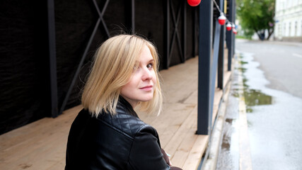 Smiling pretty blond young woman in black leather jacket and red shirt sitting outdoor on the street in the city. Spending time walking and having rest concept