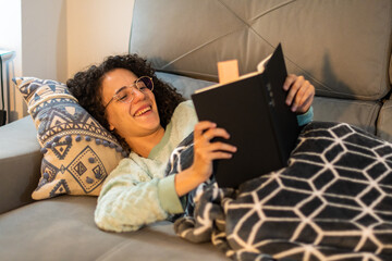 Stock photo of a beautiful and smiling young woman with curly hair and glasses reading a book at home, sitting on the coach. Curly girl with soft blanket and winter pajama reading and laughing.