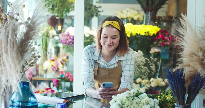 Good-looking young female worker in apron working in flower store. Charming Caucasian woman florist typing, sending message to friend on smartphone and smiling. Social media, Internet.