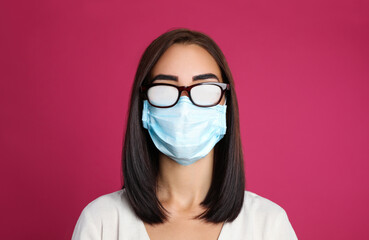Young woman with foggy glasses caused by wearing disposable mask on pink background. Protective...