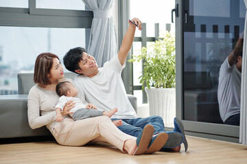 Smiling handsome Asian young man taking selfie with his wife and little daughter when they are sitting on the floor