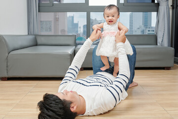 Young father lying on the floor and playing with adorable happy little girl dancing on his belly