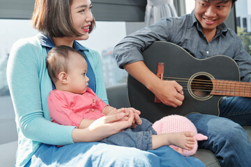 Serious little baby girl sitting on laps of her mom and lookng at her dad playing guitar and singing