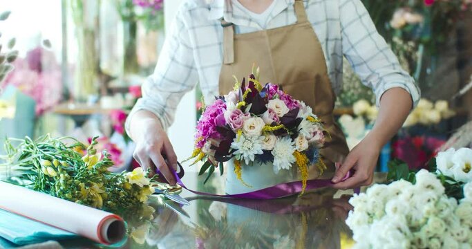 Close up of charming young Caucasian florist wearing apron, making bouquet. Beautiful female flower seller decorating, tying purple ribbon around hatbox with blossoms. Business, nature concept.