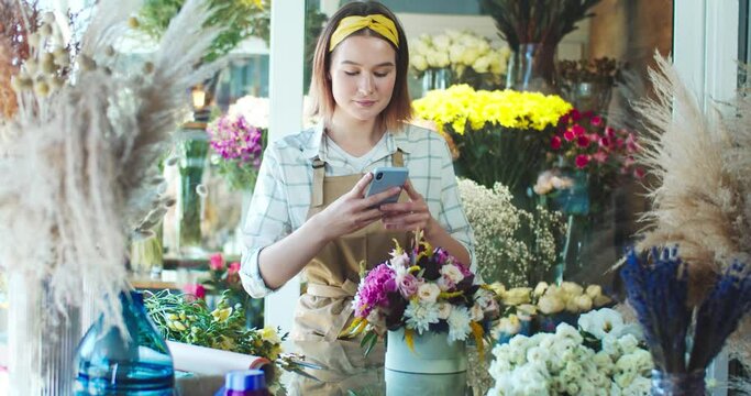 Cheerful Caucasian flower shop seller wearing apron, working in store. Pretty young female florist taking picture for customer and sending photo to client. E-commerce, business, online concept.