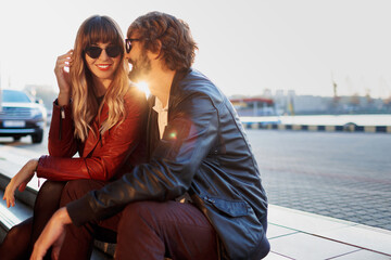 Outdoor fashion image of  stylish couple in casual outfit, leather jacket and sunglasses sitting on...