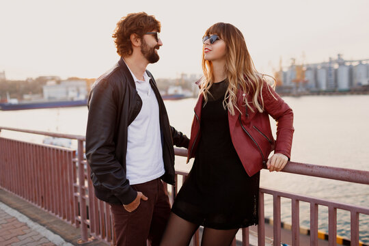 Outdoor fashion image of  stylish couple in casual outfit, leather jacket and sunglasses standing on the bridge.   Handsome man with beard with his girlfriend spending romantic time together.