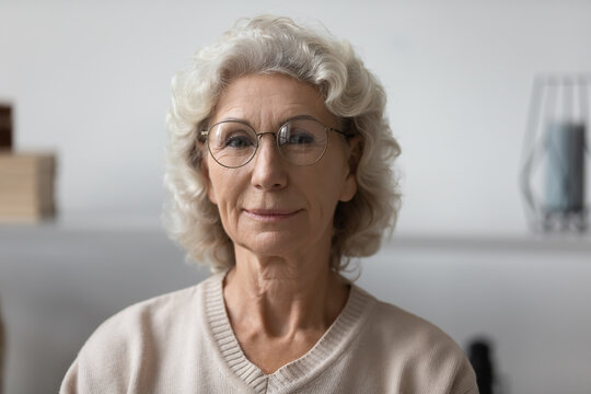 Head shot portrait confident mature woman wearing glasses at home, beautiful middle aged senior female with grey hair posing for photo in living room, sitting on couch, coach shooting vlog