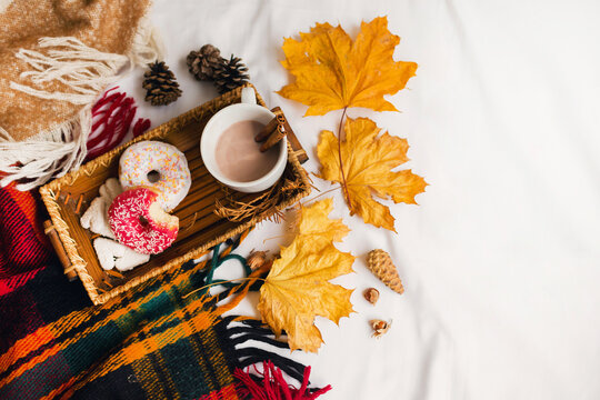  cozy autumn image of tasty breakfast in bed on wooden tray with cup of cacao, cinnamon, cookies and glazed donuts.Warm toned colors, top view, yellow leaves and cones.