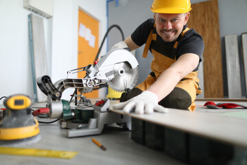Master man in yellow helmet smiles and cuts laminate board with machine. Renovation services concept