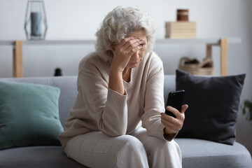 Unhappy stressed mature middle aged woman looking at phone screen, sitting on couch in living room, upset frustrated senior female reading bad news in message or social network, problem with device