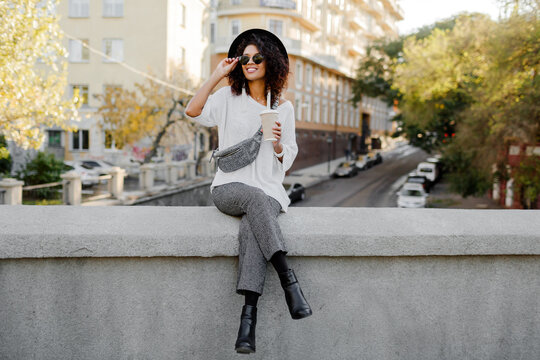 Outdoor positive image of smiling pretty black woman in white sweater and black hat  enjoying   coffee to go.  Urban background.