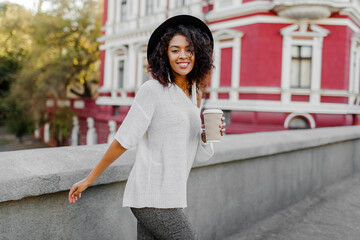 Playful woman wraps herself over her shoulder and looking in camera. Wearing. Stylish hipster outfit. Afro hairstyle . Perfect spring  leisure time in the evening City.