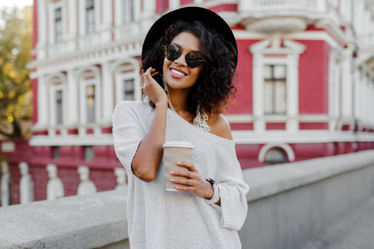 Soft toned lifestyle outdoor image of blissful black woman walking in spring  city with cup of cappuccino or hot tea. Hipster outfit. Oversize white sweater, black hat, stylish accessories.