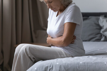 Close up frustrated mature senior woman touching belly, feeling pain and discomfort, sitting on bed at home, unhappy upset middle aged female suffering from stomachache, food poisoning, gastritis