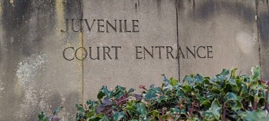 Weathered stone slab sign with the words juvenile Court Entrance engraved into it with foliage at...