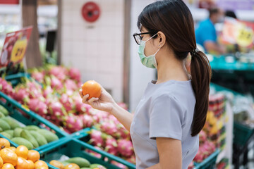 Asian woman wearing surgical face mask and holding orange fruit in supermarket or grocery, protect coronavirus inflection. Hygiene, new normal and life under covid-19 pandemic