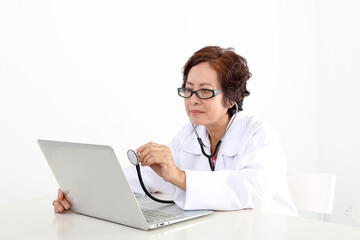 Elderly asian Chinese female doctor on white background wearing white apron checking diagnostics on notebook laptop pc with stethoscope
