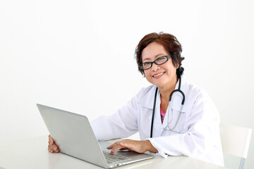 Elderly asian Chinese female doctor on white background wearing white apron stethoscope look at camera typing working at laptop notebook pc