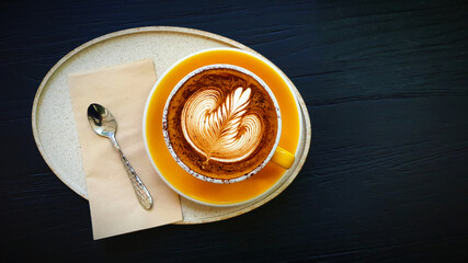 Top view of cup of art cappuccino or latte coffee putting in tray with tea spoon and napkin on dark blue wooden background. Flat lay of hot drinking with copy space on right. Food design concept.