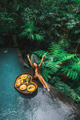 Happy girl relaxing and eating floating breakfast in jungle pool on luxury villa in Bali....