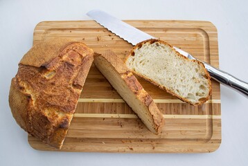 Food composition in natural light. Crispy homemade bread (French roll), sliced on a wooden board. Nearby is a fragment of a metal knife
