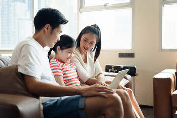 Obraz na płótnie Canvas Happy Asian family spending time together at home with laptop. Lifestyle young Asian mother father and daughter have relaxing time together while watching tv from online internet. 