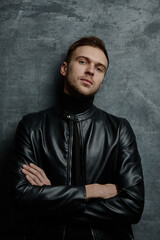 Caucasian unshaven sexy male model with brutal serious face in black leather jacket with crossed arms looking at camera on dark background isolated