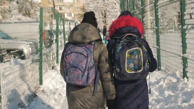Children coming home from school on a winter afternoon. Rear view of two teenage girls with backpacks walking on a snowy street home from school.