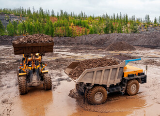 A wheel loader in a mountainous forested area loads a mining truck with a bucket, dumping the mountain soil into it. Gold mining industry. © Letopisec