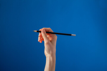 Hand Holding Black Pencil On Blue Background Photo A hand gently holds up a pencil in front of a bright red background, gesturing a pencil stroke.