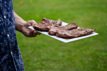 Male hand in a blue shirt holding a white plate with grilled beef steaks. Green grass background.