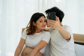 young Asian pregnant woman with husband on the white bed in the house taking selfie  with mobile cellphone