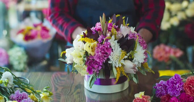 Attractive young male florist working in modern flower store. Handsome Caucasian man decorating bouquet with purple ribbon around hatbox in shop. Decor, business, commerce concept.