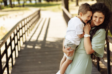 Fototapeta premium Portrait of adorable mother with overjoyed son at the park, beautiful woman hold in arms little boy, smiling, hugging, family time, motherhood and childhood concept