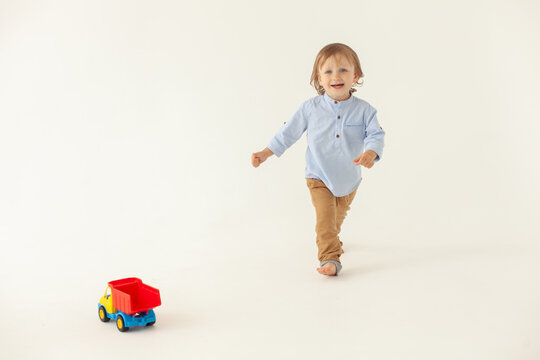 A 2 Year Old Boy In A Blue Shirt And Beige Trousers Runs And Laughs On A White Background Next To Him A Toy Car