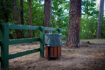 Clean and neat rubbish bin in forest. Green wooden fence next to. Rest and leisure area for tourists with trash bin.