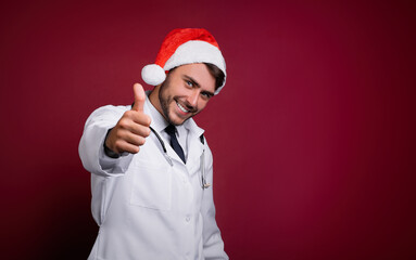 Young handsome doctor in white uniforme and Santa Claus hat standing in studio on red background smile and finger in camera