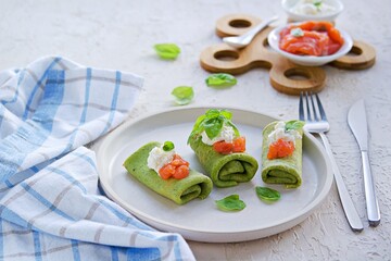 Spinach pancakes rolled up on a white plate on a light concrete background. Served with salted salmon and soft cheese. Spinach recipes.