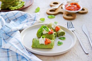 Spinach pancakes rolled up on a white plate on a light concrete background. Served with salted salmon and soft cheese.
