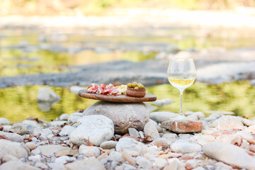 appetizing delicious snacks of meat and cheese on a wooden board and a glass of wine stand on the river bank at a picnic