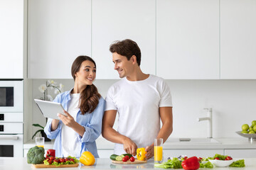 Cute couple cooking in kitchen, man chopping vegetables while his girlfriend is using a digital tablet looking for a recipe. Family enjoying together.