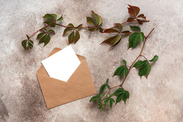 Autumn stationery scene, blank card in craft envelope and branch with wild grape leaves. Beige grunge background. Wedding invitation mockup. Corporate identity design. Top view, flat lay.